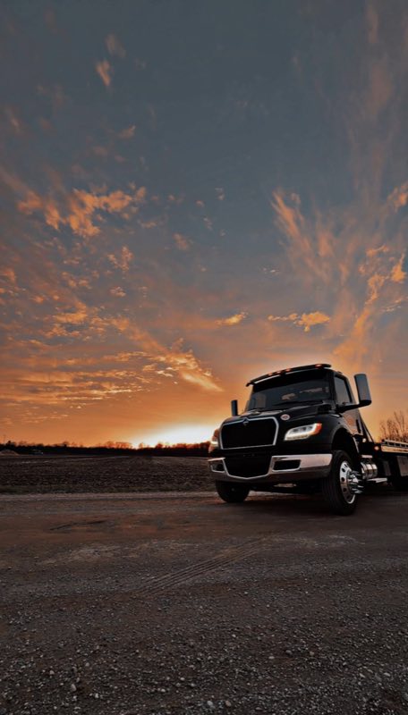 RR Towing & Recovery flatbed tow truck at sunset, headlights on, parked on a gravel lot